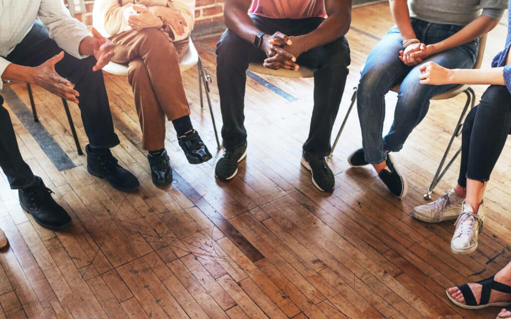 Several individuals seated on chairs arranged in a circle, visible from the waist down, gesture and hold notepads suggesting an informal collaborative meeting.