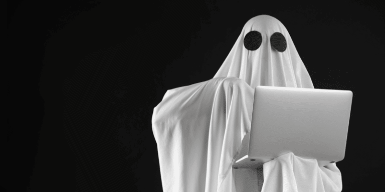 A figure draped in a white sheet with eye holes, ghost-like, stands against black, holding an open laptop—symbolizing ghost users.