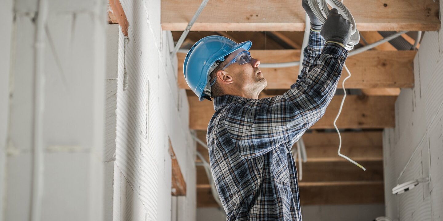 A construction worker, wearing a blue hard hat and plaid shirt, stands on a ladder while installing wiring on a ceiling. The worker is focused and uses both hands to securely position the wires. Dim lighting and exposed beams suggest an unfinished or industrial setting. The scene conveys precision and safety in a construction environment.